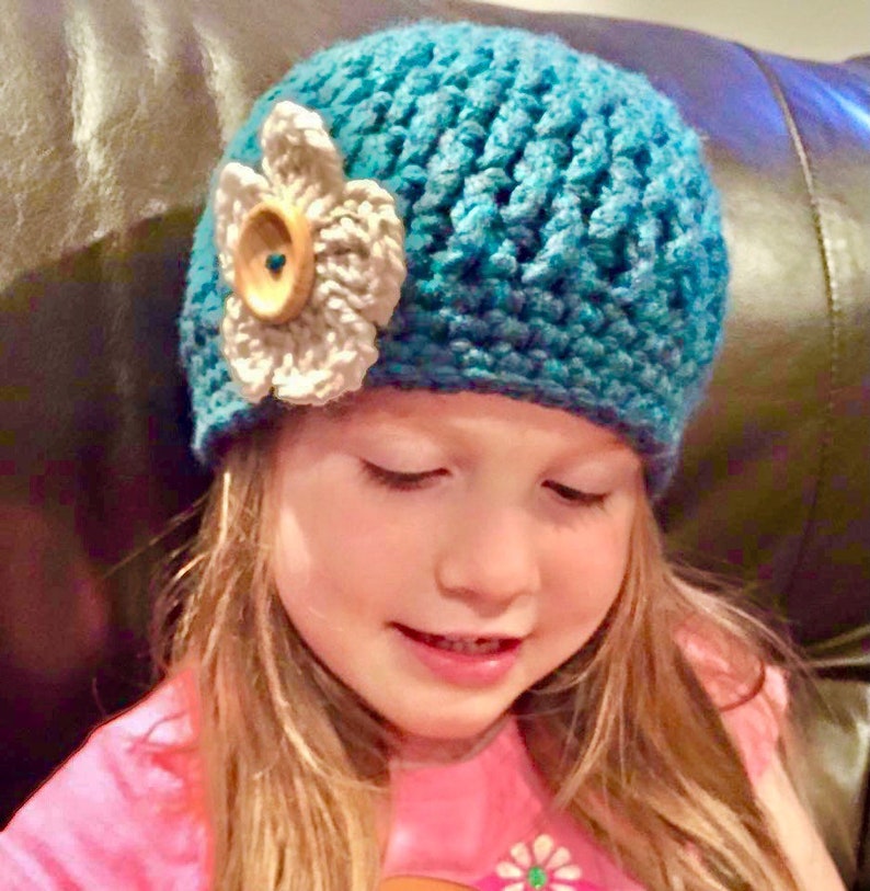 May include: A young girl wearing a teal blue crocheted hat with a white crocheted flower and a brown button. The hat is worn on her head and she is looking down.