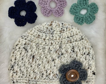May include: A white crocheted beanie with a brown button and a gray flower accent. The beanie has a speckled pattern and is surrounded by three crocheted flowers in different colors.