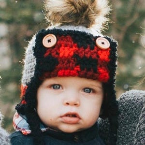 May include: A baby wearing a handmade winter hat with a red and black plaid pattern. The hat has ear flaps, wooden button accents, and a fluffy brown pom-pom on top. The baby is wearing a dark blue shirt.