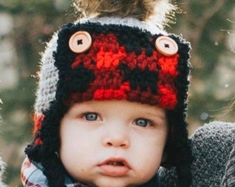 May include: A baby wearing a handmade winter hat with a red and black plaid pattern. The hat has ear flaps, wooden button accents, and a fluffy brown pom-pom on top. The baby is wearing a dark blue shirt.
