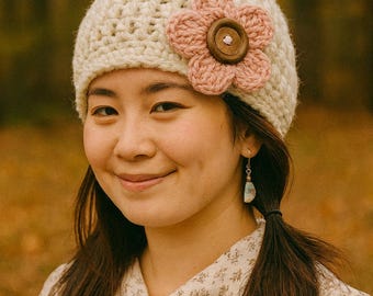 May include: A cream-colored crocheted beanie featuring a large, five-petaled pink crocheted flower attached to the side. The center of the flower is adorned with a round, dark brown wooden button with four holes in the middle.