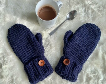 May include: A pair of navy blue crochet mittens with a textured stitch pattern. Each mitten features a cuff with a decorative wooden button. A white mug of hot chocolate and a spoon are in the background.