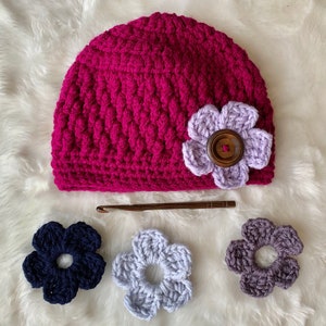 May include: A pink crocheted beanie with a purple flower and a brown button. The beanie is on a white fluffy surface with a crochet hook and two other crocheted flowers.