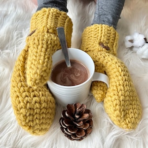 May include: A pair of yellow crocheted mittens with a button detail, held by a person, with a mug of hot chocolate and a pine cone on a white fuzzy surface.