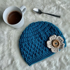 May include: A teal blue crochet beanie with a beige flower and wooden button accent. A white mug with a dark brown beverage and a silver spoon are also in the image. The beanie is a handmade item.