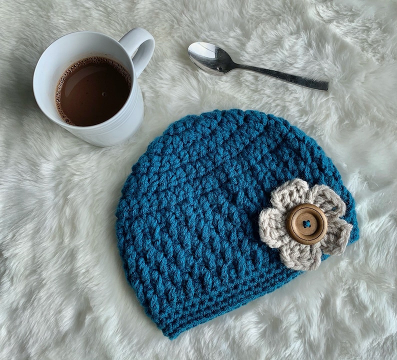 May include: A teal blue crocheted beanie with a white crocheted flower and a brown button in the center. The beanie is on a white fuzzy surface.