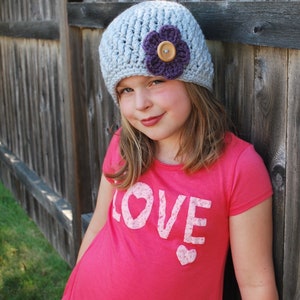 May include: A young person wearing a gray crocheted hat with a purple flower and a pink shirt that says "LOVE" in white letters.