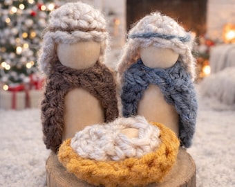 May include: Handmade Christmas nativity scene featuring two figures with knitted shawls and head coverings. A knitted manger sits on a wooden base. The figures are set against a backdrop of a Christmas tree and fireplace.