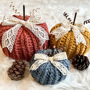 May include: Three knitted pumpkins with lace accents. The pumpkins are in shades of red, yellow, and blue. The pumpkins are sitting on a white fluffy surface.