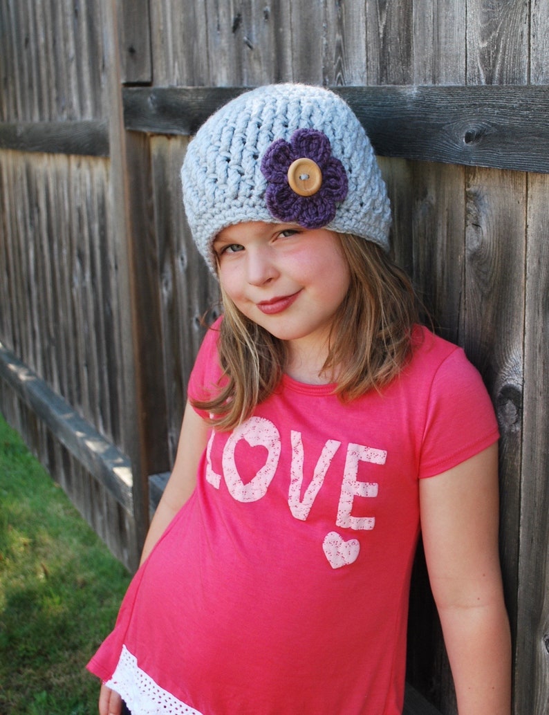May include: A young person wearing a gray crocheted hat with a purple flower and a pink shirt that says "LOVE" in white letters.