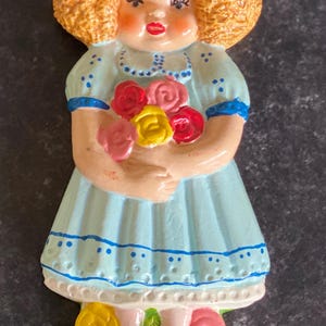 May include: A ceramic figurine of a girl with curly blonde hair wearing a blue dress with white polka dots and holding a bouquet of pink, red, and yellow roses. She is standing on a green base with yellow and pink roses.