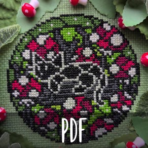 May include: A cross-stitch pattern with a floral design in shades of black, gray, green, and red. The word "PDF" is displayed in white. The design is surrounded by green leaves and small red and white mushroom decorations.