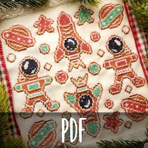 Pode incluir: Padrão de ponto cruz com desenhos de biscoitos de gengibre com tema espacial. O design inclui um foguete, astronautas, planetas e estrelas em tons de vermelho, verde e castanho. A palavra "PDF" é exibida na parte inferior da imagem.