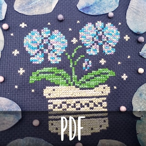 Puede incluir: Patrón de punto de cruz de una planta en maceta con flores azules y moradas sobre un fondo azul oscuro. El patrón está rodeado de pétalos de flores azules y cuentas blancas.