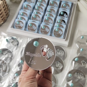 May include: A silver round mirror with a blue pearl in the centre. The mirror has a cutout of two baby footprints and the text "Hosgeldin Isa" engraved on it.