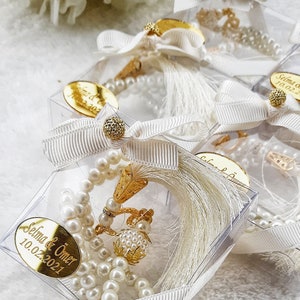 May include: White pearl prayer beads with a gold tassel and a gold charm in a clear plastic box with a white ribbon and a gold tag that says "Selma & &Ouml;mer 10.02.2021".