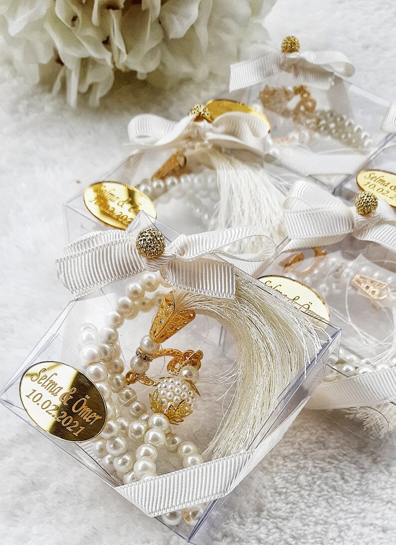 May include: White pearl prayer beads with a gold tassel and a gold charm in a clear plastic box with a white ribbon and a gold tag that says "Selma & &Ouml;mer 10.02.2021".