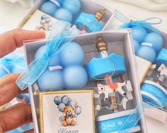 May include: A blue and white favour box with a blue and white carousel, a blue balloon candle, and a small card with a teddy bear and balloons. The card says "Kenan 1 Yasinda".