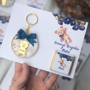 May include: A white and gold keychain with a blue bow and a teddy bear charm. The keychain is attached to a gold key ring. The keychain is on a white card with the text "Aramıza Hoşgeldin Mahir" and a chocolate bar with a teddy bear design and the text "Mahir".
