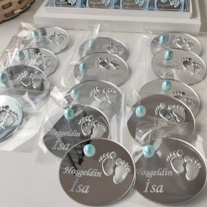 May include: Silver oval-shaped mirror favours with a baby footprint design and the text "Hoşgeldin Isa" in a cursive font. Each favour has a light blue pearl embellishment.