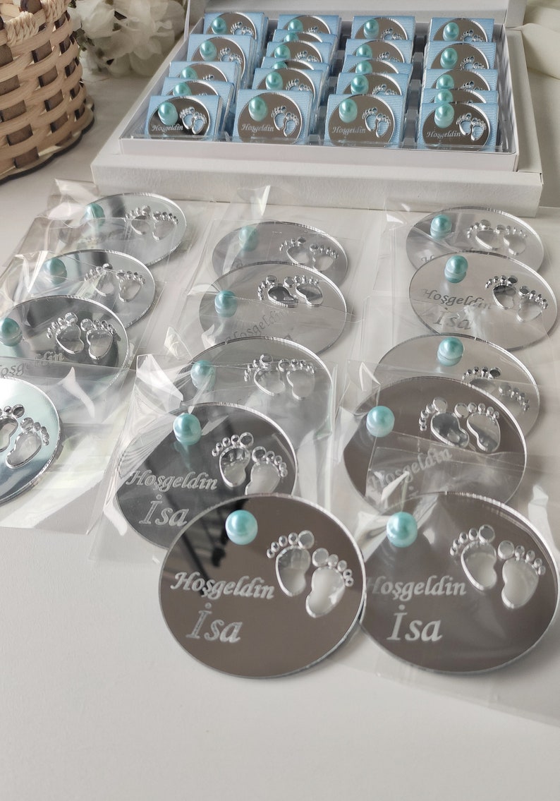 May include: Silver oval-shaped mirror favours with a baby footprint design and the text "Hoşgeldin Isa" in a cursive font. Each favour has a light blue pearl embellishment.