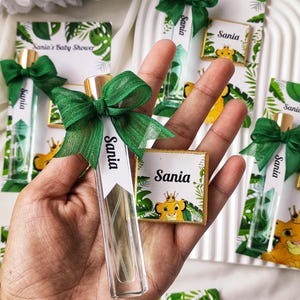 Puede incluir: Frasco de perfume de vidrio transparente con tapón dorado, lazo verde y etiqueta que dice "Sania". También se ve una pequeña tableta de chocolate con un dibujo de león y el nombre "Sania". El tema es un baby shower.