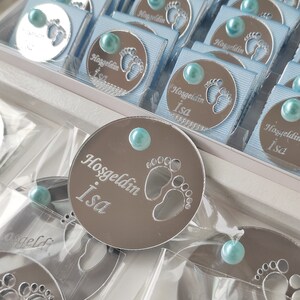 May include: A collection of small, round, silver-coloured favours with a blue ribbon and a blue pearl. The favours have a design of a baby's footprint and the text "Hosgeldin Isa".
