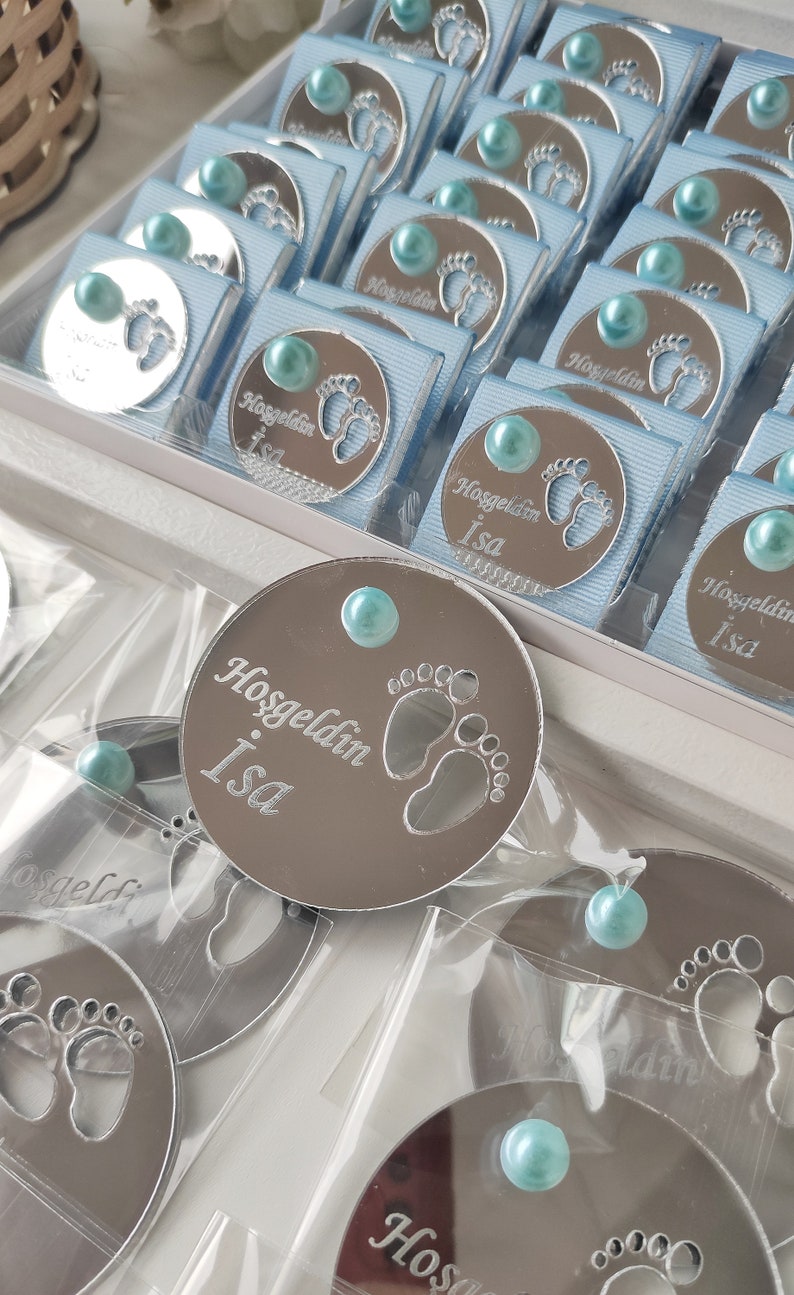 May include: A collection of small, round, silver-coloured favours with a blue ribbon and a blue pearl. The favours have a design of a baby's footprint and the text "Hosgeldin Isa".