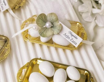 May include: Gold-colored trays filled with white candies, some adorned with floral decorations and tags that read "Hoşgeldin Arjin Zana". The trays are set on a white, textured surface, with a woven basket and white flowers in the background.