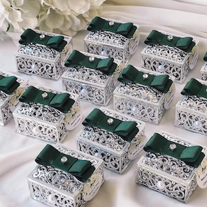 Puede incluir: Varias cajas de regalo plateadas con diseños intrincados y lazos de satén verde. Cada caja tiene una pequeña etiqueta blanca con el texto "Boda de [Nombre] & [Nombre] [Fecha]" impreso en ella.