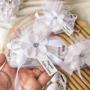 Puede incluir: Favores adornados con flores blancas con un borde plateado, que contienen caramelos blancos, se presentan en una bandeja de madera. Cada favor está decorado con una flor blanca y una etiqueta que dice "Hoşgeldin Arjin Zana".