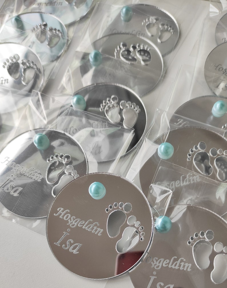 May include: Silver round mirror with a blue gem in the centre. The mirror has a design of two baby footprints and the text "Hosgeldin Isa".