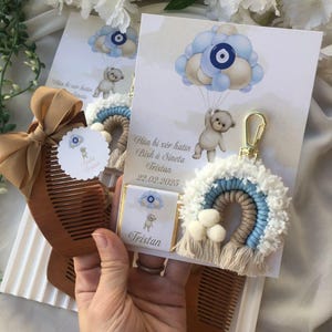 Puede incluir: Recuerdos de baby shower, que incluyen un peine de madera con un lazo, un llavero de arcoíris de macramé y tarjetas con un diseño de oso de peluche y globos. Los recuerdos son en tonos marrón, azul y blanco.