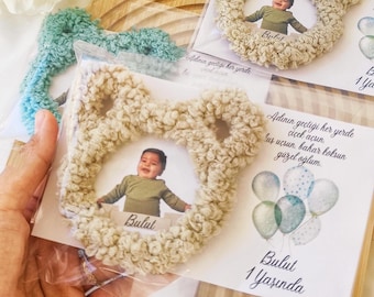May include: A beige fuzzy bear-shaped frame with a photo of a baby boy inside. The frame is on a white card with the text "Bulut 1 Yaşında" and a blue and green balloon design. The card is on a wooden surface.