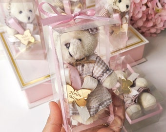 May include: A set of five clear plastic boxes with pink ribbon bows, each containing a small white teddy bear keychain. The teddy bears are wearing plaid dresses and have a gold butterfly charm with the inscription "Helin Nour".