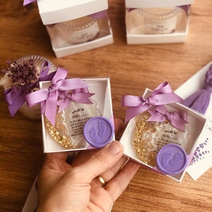May include: Two small white boxes with purple ribbon bows, each containing a clear glass jar with gold flakes and a purple wax seal with a footprint design. The boxes are labeled with the Arabic text "Ayah al-Kursi" which translates to "Verse of the Throne".