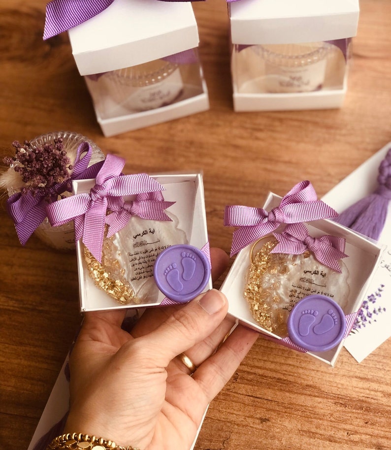May include: Two small white boxes with purple ribbon bows, each containing a clear glass jar with gold flakes and a purple wax seal with a footprint design. The boxes are labeled with the Arabic text "Ayah al-Kursi" which translates to "Verse of the Throne".