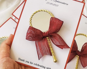 May include: A white card with red trim and gold lettering that says "Suheyri's Quinceanera 25/10/2025" and "Thank you for celebrating with us!" A small gold hand mirror with a red bow is attached to the card.
