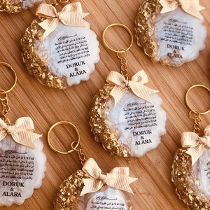 May include: A set of six gold and white keychains with a white background and gold flecks. Each keychain features a white circle with Arabic text and the names "DORUK & ALARA" printed in black. The keychains are attached to gold key rings with a gold bow.