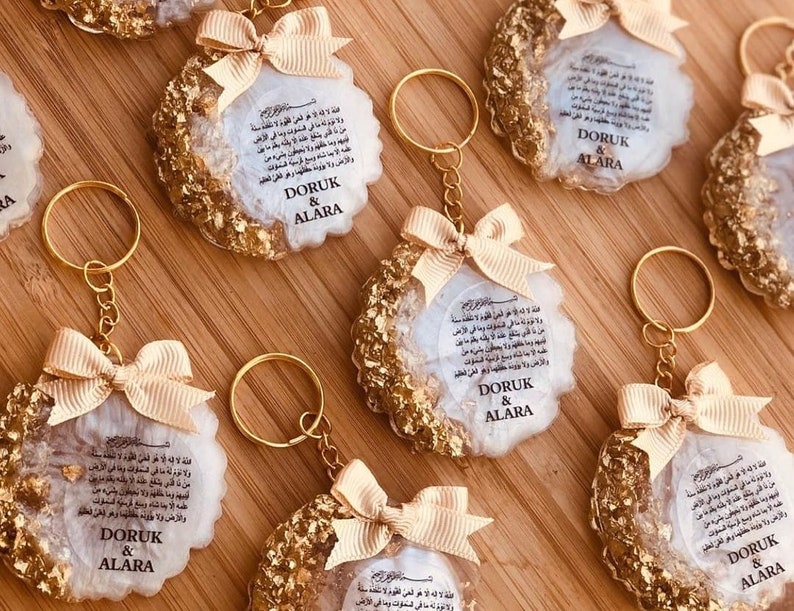 May include: A set of six gold and white keychains with a white background and gold flecks. Each keychain features a white circle with Arabic text and the names "DORUK & ALARA" printed in black. The keychains are attached to gold key rings with a gold bow.