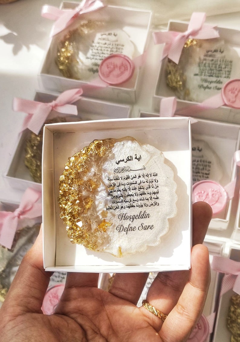 May include: A white box with a pink ribbon contains a white and gold resin ornament with Arabic text and the words "Hoşgeldin Defne Sare".