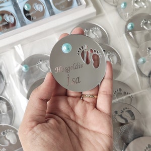 May include: A silver round mirror with a blue gem in the centre. The mirror has a cutout of two baby footprints and the text "Hosgeldin Isa" in silver lettering.