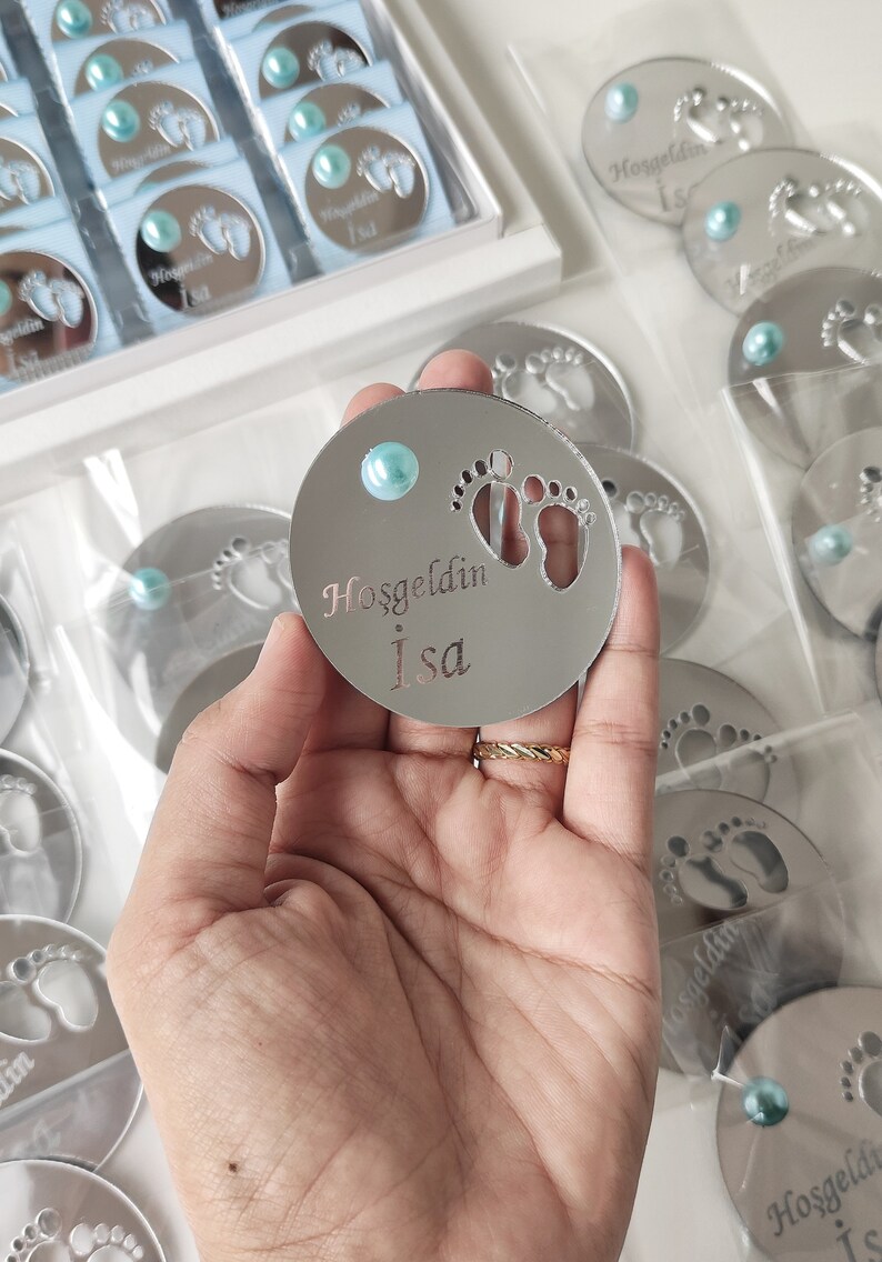 May include: A silver round mirror with a blue gem in the centre. The mirror has a cutout of two baby footprints and the text "Hosgeldin Isa" in silver lettering.