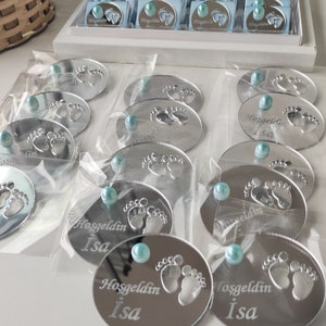 May include: Silver mirror baby foot-shaped favours with a blue pearl and the text "Hosgeldin Isa" engraved on each.