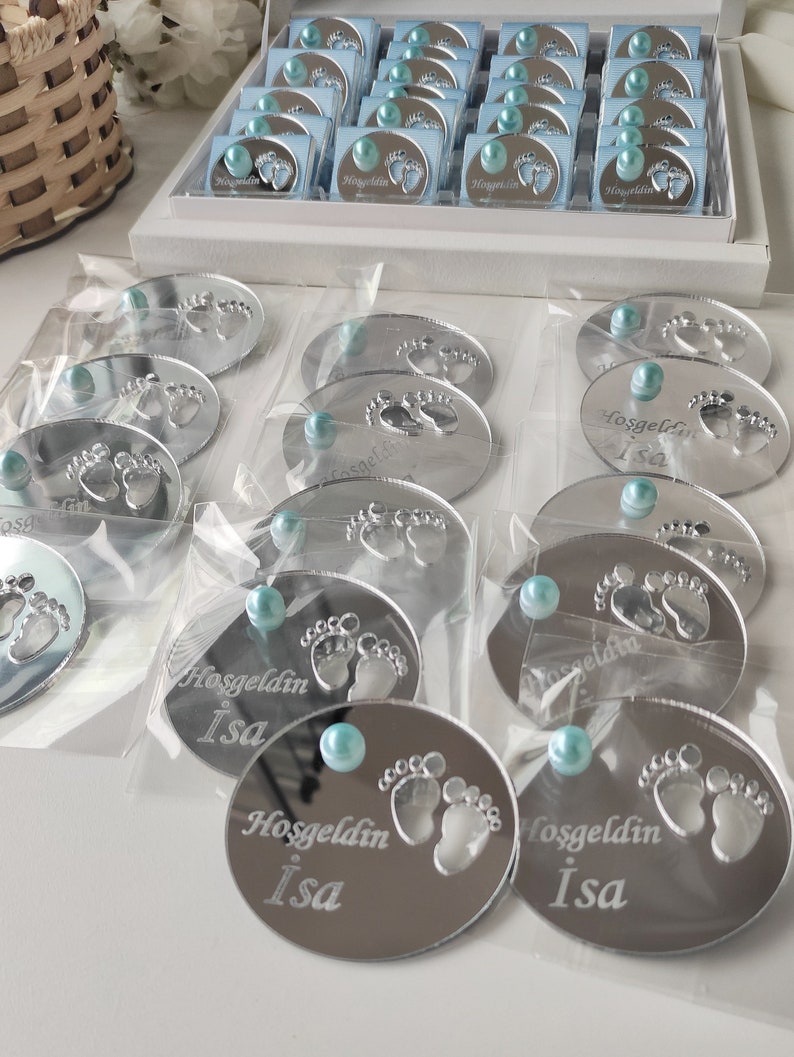 May include: Silver mirror baby foot-shaped favours with a blue pearl and the text "Hosgeldin Isa" engraved on each.