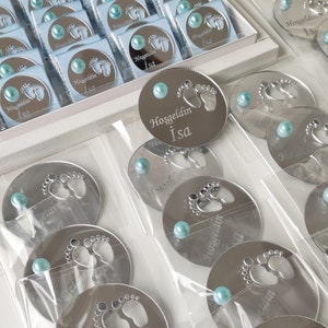 May include: Silver round mirror favours with a baby footprint design and the text "Hoşgeldin Isa" in white. Each favour has a light blue pearl attached.