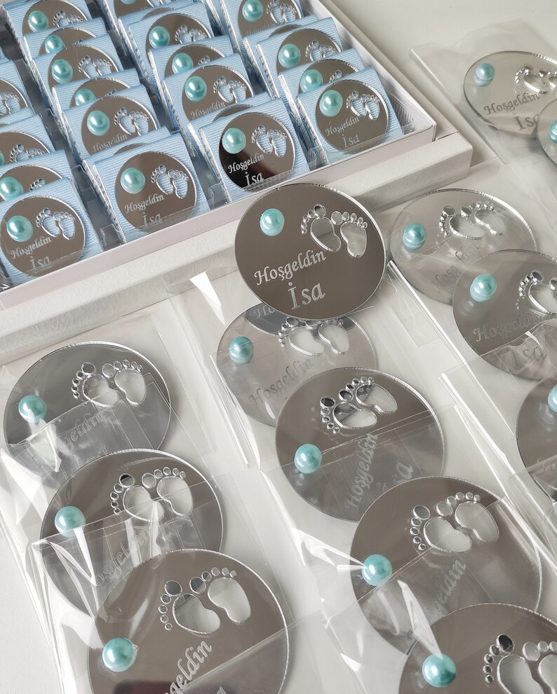 May include: Silver round mirror favours with a baby footprint design and the text "Hoşgeldin Isa" in white. Each favour has a light blue pearl attached.
