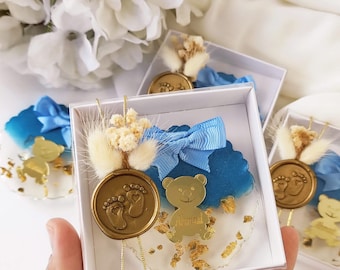 May include: A small white box with a clear lid contains a blue and white baby shower favor. The favor includes a blue and white wax seal with a footprint design, a gold teddy bear charm with the name "Ahmed" on it, and a small bouquet of dried white flowers.