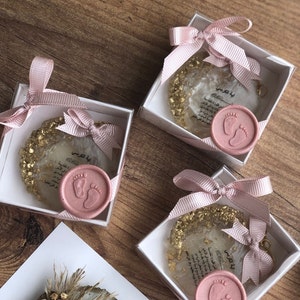 May include: Three small white boxes with pink ribbon bows and gold glitter inside. Each box contains a clear resin keychain with gold flakes and a pink wax seal with a baby footprint design. The keychain has the Arabic text "Ayah al-Kursi" printed on it.