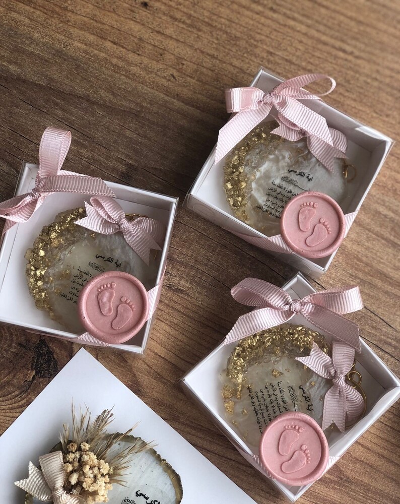 May include: Three small white boxes with pink ribbon bows and gold glitter inside. Each box contains a clear resin keychain with gold flakes and a pink wax seal with a baby footprint design. The keychain has the Arabic text "Ayah al-Kursi" printed on it.